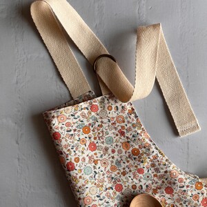 May include: An apron featuring a dense floral pattern with small red, orange, and light blue flowers on a cream background. The apron has wide, beige herringbone-patterned fabric straps and a dark metal ring adjuster. A wooden spoon rests on the fabric.