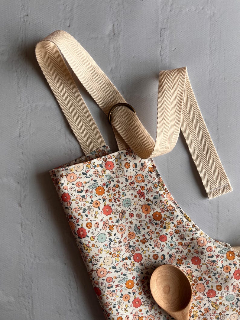 May include: An apron featuring a dense floral pattern with small red, orange, and light blue flowers on a cream background. The apron has wide, beige herringbone-patterned fabric straps and a dark metal ring adjuster. A wooden spoon rests on the fabric.
