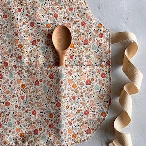 May include: A kitchen apron featuring a dense floral pattern in shades of coral, orange, and light blue on a cream background. The apron includes a front pocket containing a smooth, light-colored wooden spoon and long, beige herringbone-weave ties.