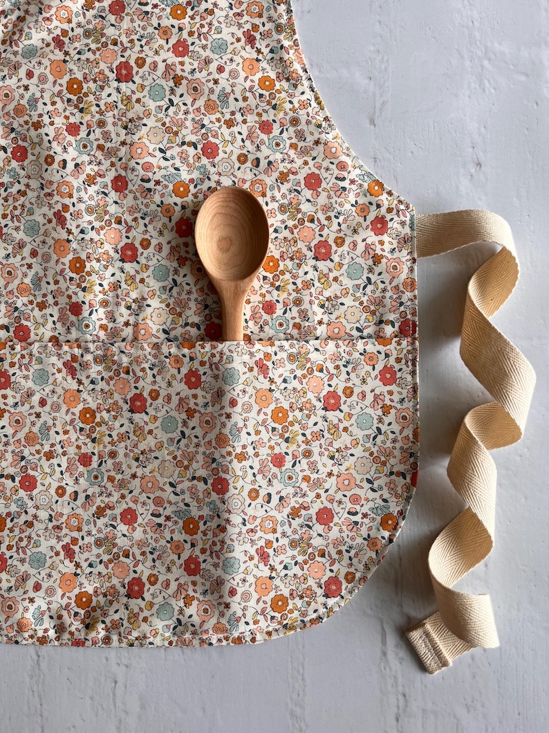 May include: A kitchen apron featuring a dense floral pattern in shades of coral, orange, and light blue on a cream background. The apron includes a front pocket containing a smooth, light-colored wooden spoon and long, beige herringbone-weave ties.