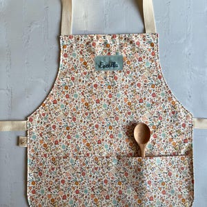 May include: A white apron with a floral print in shades of pink, orange, and blue. The apron has a large pocket on the front and a name tag that says "Estella".