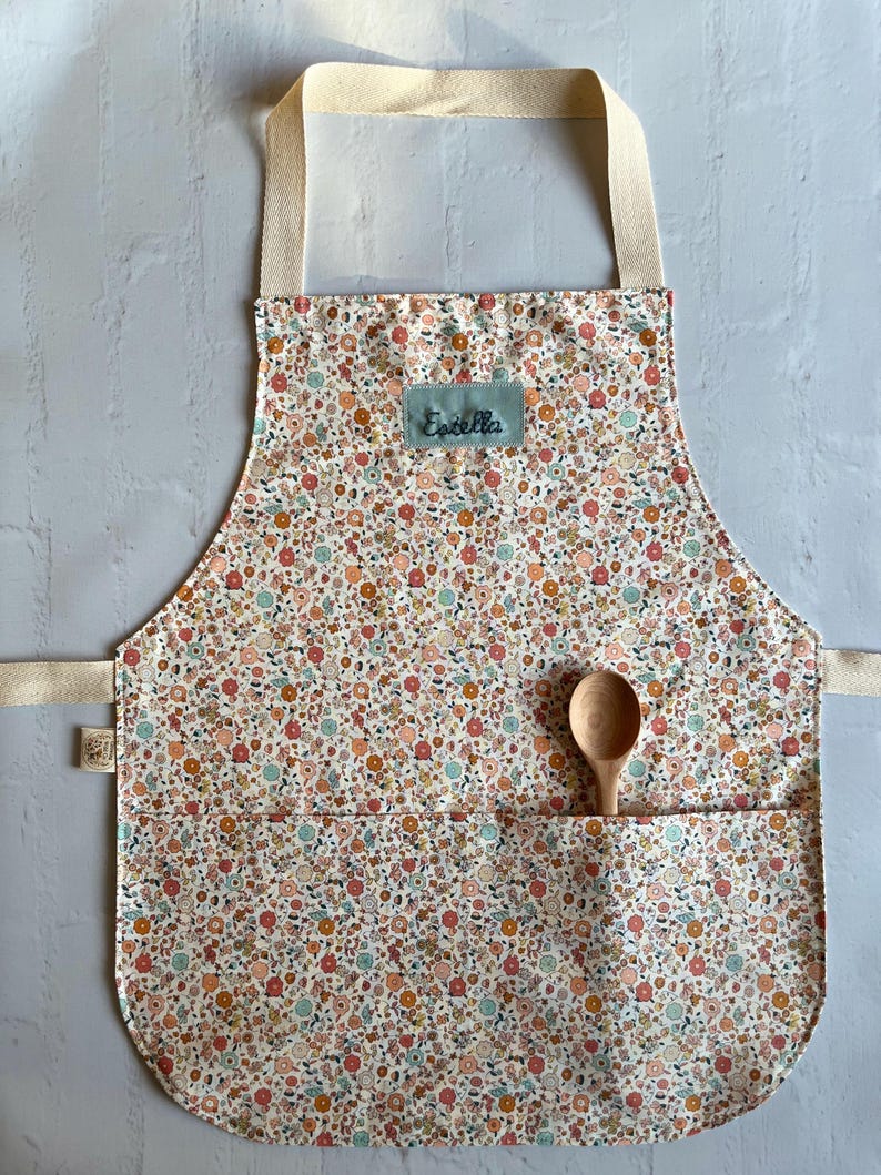 May include: A white apron with a floral print in shades of pink, orange, and blue. The apron has a large pocket on the front and a name tag that says "Estella".