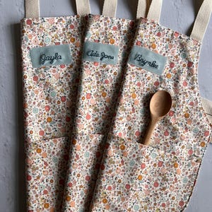 May include: Three floral print aprons with cream-colored straps and pockets. Each apron has a light blue patch with a name embroidered on it: Jayla, Ada Jane, and Brynlee. A wooden spoon rests in one pocket.