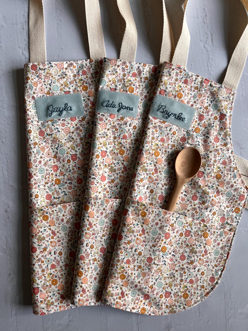 May include: Three floral print aprons with cream-colored straps and pockets. Each apron has a light blue patch with a name embroidered on it: Jayla, Ada Jane, and Brynlee. A wooden spoon rests in one pocket.