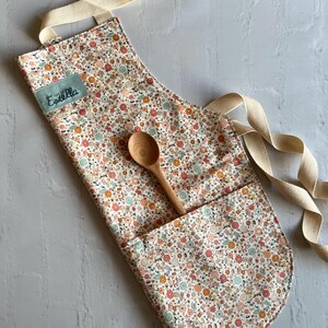 May include: A white apron with a floral print in shades of orange, pink, and green. The apron has a large pocket on the front and a wooden spoon is resting in the pocket. The apron has a cream-colored strap and the name "Estella" is embroidered on the front.