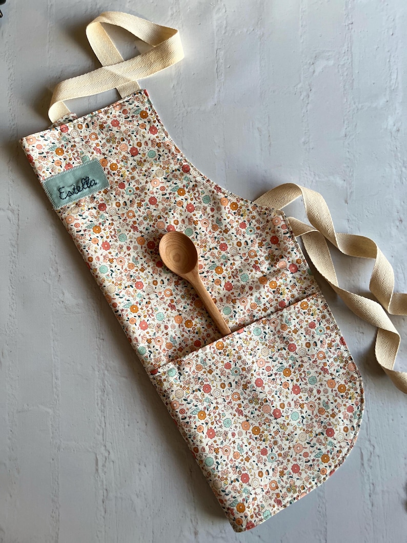 May include: A white apron with a floral print in shades of orange, pink, and green. The apron has a large pocket on the front and a wooden spoon is resting in the pocket. The apron has a cream-colored strap and the name "Estella" is embroidered on the front.
