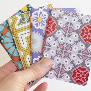 May include: Four small notebooks with colourful patterned covers. The covers feature geometric designs in shades of blue, yellow, red, and purple.
