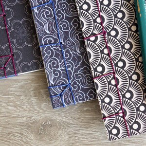 May include: Three handmade journals with black covers and different patterned designs. The covers are bound with a dark purple thread. A green pencil is lying on top of the journals.