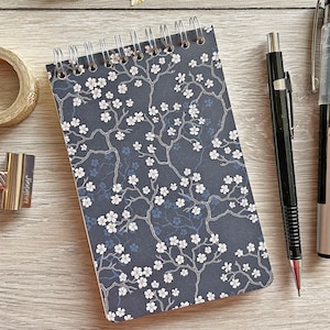 May include: A small, spiral bound notebook with a dark blue cover featuring a white floral pattern. The notebook is lying on a wooden surface with two pens beside it.
