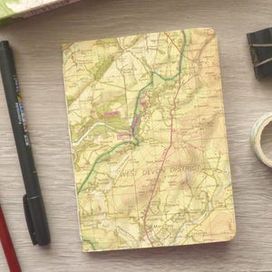 May include: A rectangular notebook with a cover featuring a vintage topographical map of the West Devon District. The notebook is accompanied by a red pencil, a black felt-tip pen, a black metal binder clip, and a roll of patterned adhesive tape.