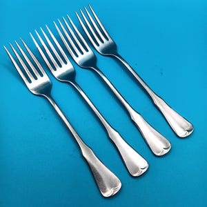 May include: A set of four matching silver-toned metal forks arranged diagonally. Each fork features a long, slender handle that widens slightly toward the base, a smooth polished finish, and four tines designed for dining and food service.