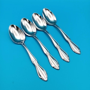 May include: A set of four polished silver-tone metal spoons arranged diagonally. Each spoon features a smooth, oval-shaped bowl and a handle decorated with a subtle floral embossed pattern at the base, reflecting light on their metallic surfaces.