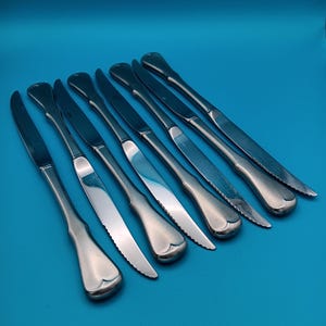 May include: A collection of eight stainless steel butter knives arranged diagonally on a blue surface. Each knife has a heart-shaped handle and a serrated edge. The blades are shiny and reflect the light.