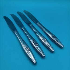 May include: Four stainless steel dinner knives with a floral rose design on the handles. The knives are arranged diagonally against a solid blue background. The blades are smooth and shiny.