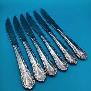 May include: Six silver-colored knives with ornate, engraved handles are arranged diagonally against a blue background. The blades are smooth and reflective. The handles feature a detailed floral and scroll design, adding a touch of elegance to the cutlery set.