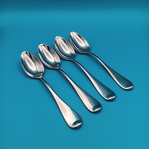 May include: Four silver spoons arranged diagonally on a solid blue background. The spoons have a classic design with elongated handles and rounded bowls. The spoons are reflective, showing the light and the background.
