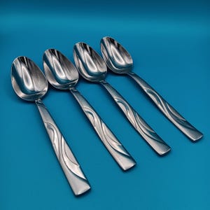 May include: Four stainless steel spoons with a reflective surface and a decorative wave pattern on the handles. The spoons are arranged diagonally against a solid blue background, showcasing their design.