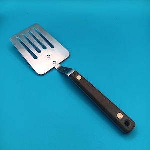 May include: A stainless steel spatula with a slotted head and a dark brown wooden handle. The handle is secured with two brass-colored rivets. The spatula is designed for flipping food while cooking.