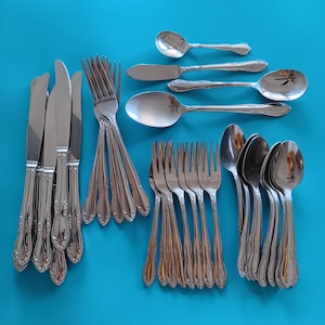 May include: A collection of stainless steel silverware, including knives, forks, and spoons, arranged on a bright blue surface. The silverware has a decorative floral pattern on the handles. The set includes various spoon sizes and butter knives.