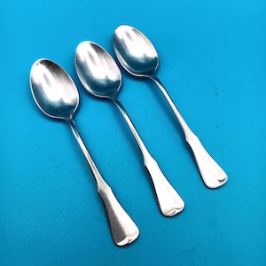 May include: Three silver-colored metal spoons arranged diagonally. Each spoon features a smooth, oval-shaped bowl and a slender handle that widens slightly toward the base, which has a rounded, flat end. The surface of the metal is reflective.