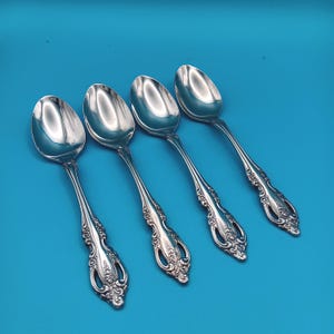May include: Four silver spoons with ornate handles and oval bowls are arranged diagonally on a blue background. The spoons have a reflective, polished surface, and the handles feature a detailed, floral design.