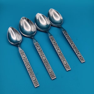 May include: Four stainless steel spoons with ornate floral designs on the handles. The spoons have a shiny, reflective surface and are arranged diagonally against a solid blue background. The handles feature a repeating pattern of flowers and geometric shapes.