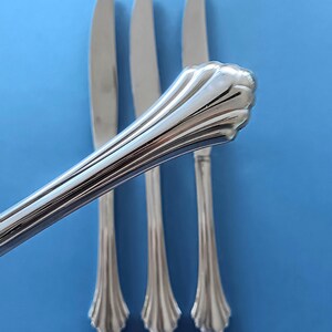 May include: Three silver dinner knives with ornate handles. The knives are arranged in a row on a blue background.