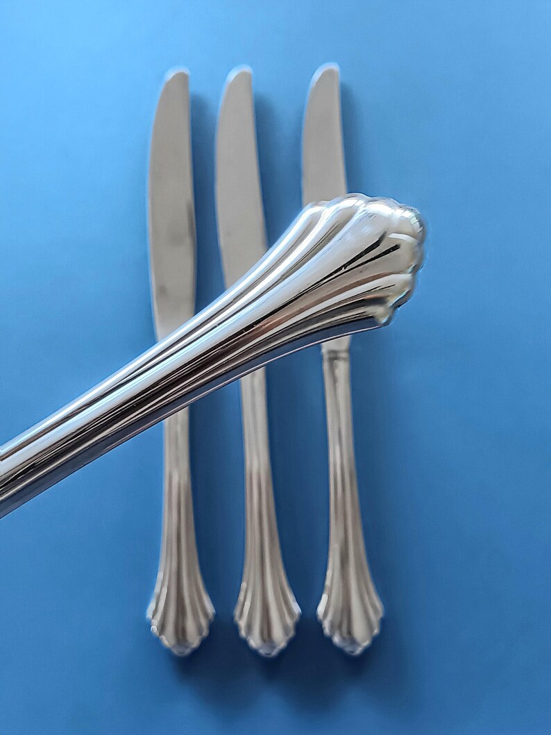 May include: Three silver dinner knives with ornate handles. The knives are arranged in a row on a blue background.