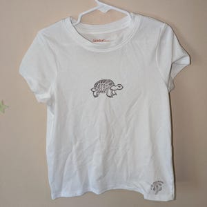 May include: White short-sleeved t-shirt with a small embroidered turtle design on the front. The shirt has a crew neck and the text "Cat & Jack" is visible on the tag.
