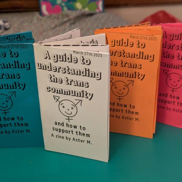 May include: A stack of five small, colorful zines titled "A guide to understanding the trans community" with a simple illustration of a person with a gender-neutral symbol. Each zine has a different color: blue, white, orange, and red. The text on the zines includes the date "March 27th 2025" and the phrase "A zine by Aster M."