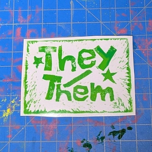May include: A white sticker with the words "They Them" printed in green, with star accents. The sticker has a textured, hand-printed appearance and is set against a blue cutting mat.