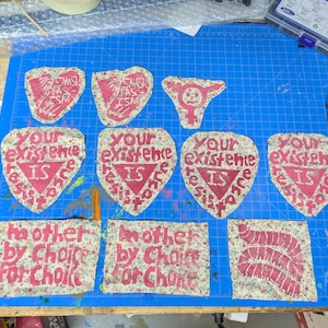 May include: A collection of fabric patches with various designs and text. Some patches feature the phrases "Your existence is resistance" and "Mother by choice for choice" in red lettering. Other patches have symbols and text.