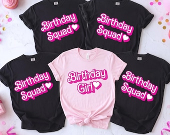 May include: Four short-sleeved t-shirts arranged on a surface. Three black shirts feature the white and pink text "Birthday Squad" with a heart. One light pink shirt in the center features the white and pink text "Birthday Girl" with a heart.