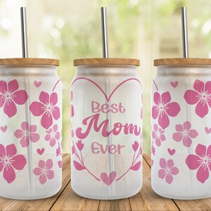 May include: Three white tumblers with pink floral designs and a straw. The center tumbler has a pink heart with the text "Best Mom Ever" inside.