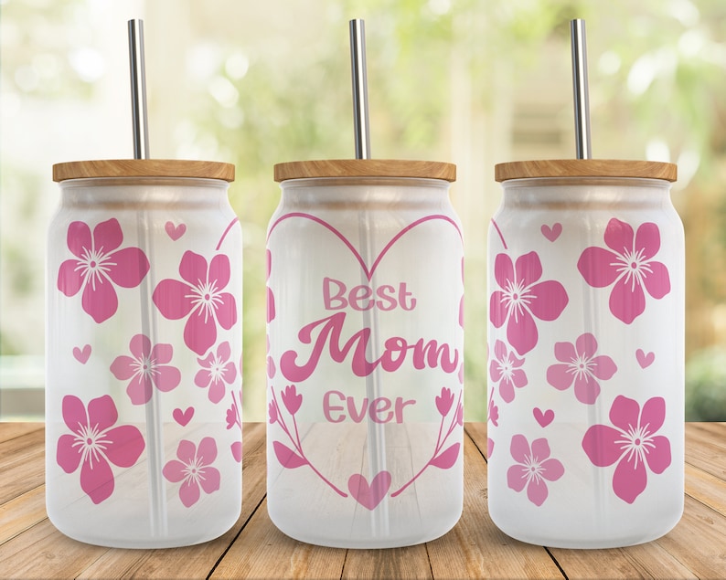 May include: Three white tumblers with pink floral designs and a straw. The center tumbler has a pink heart with the text "Best Mom Ever" inside.