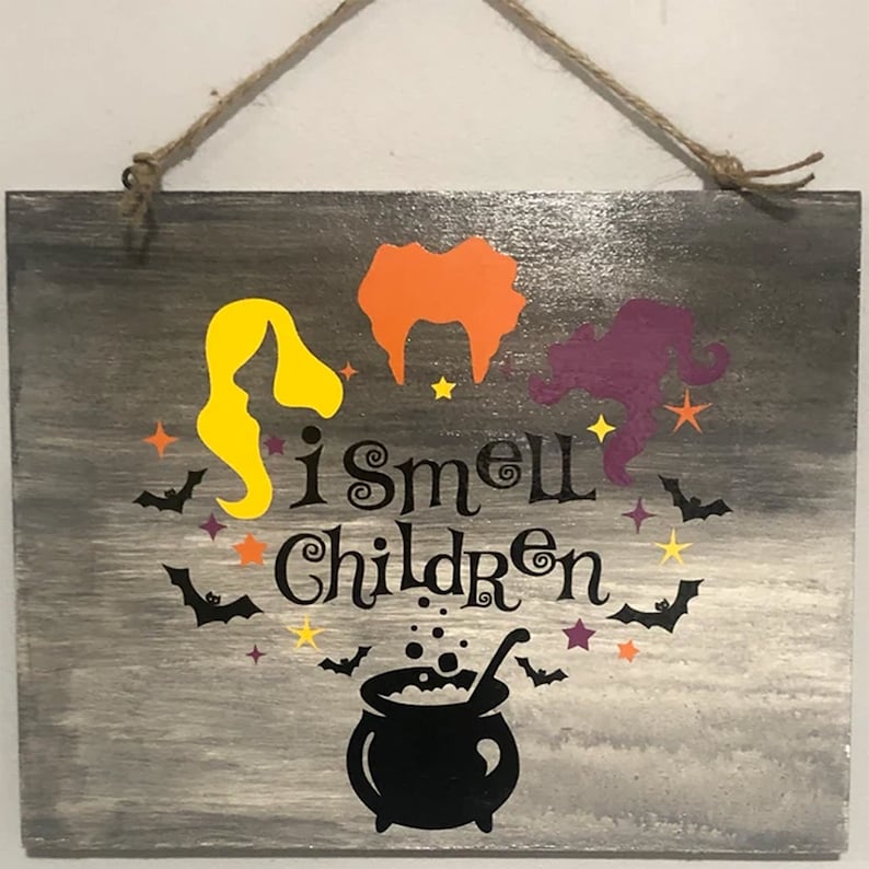 May include: A wooden sign with a gray background and a black silhouette of a cauldron with steam coming out of it. The sign says "I smell children" with three witches in the background. The witches are yellow, orange, and purple.