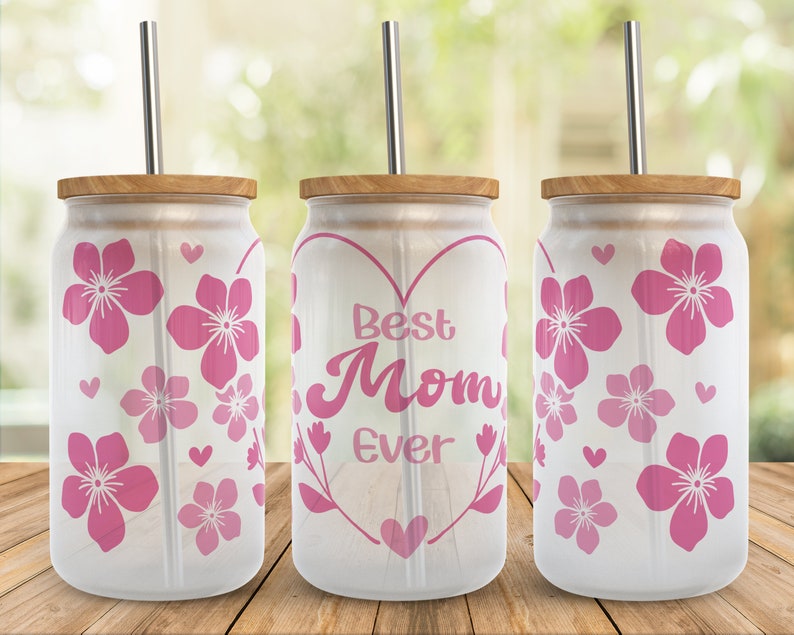 May include: Three white tumblers with pink floral designs and a straw. The center tumbler has a pink heart with the text "Best Mom Ever" inside.