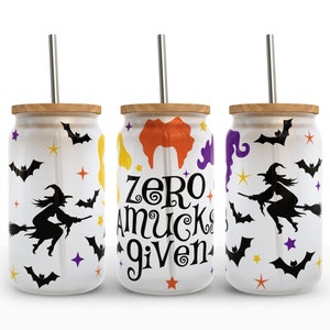以下が含まれることがあります： 黒い魔女、コウモリ、星が描かれた白いハロウィンテーマのタンブラーが3つ。真ん中のタンブラーには「zero mucks given」の文字が書かれています。