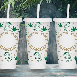 May include: Three white plastic tumblers with lids and straws. Each tumbler is decorated with green cannabis leaves, a gingerbread man wearing a Santa hat, and the text "LET'S GET BAKED" in gold lettering. The tumblers are on a wooden surface.