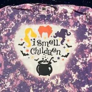 May include: Purple and white tie-dye t-shirt with a white heart-shaped design. The design features three witches, a cauldron, bats, stars, and the text "I smell children".