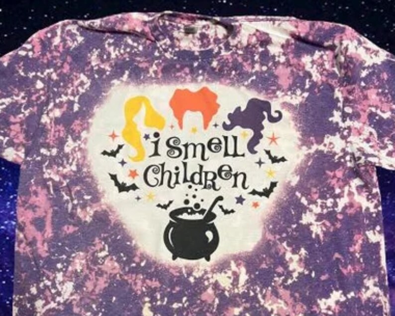 May include: Purple and white tie-dye t-shirt with a white heart-shaped design. The design features three witches, a cauldron, bats, stars, and the text "I smell children".