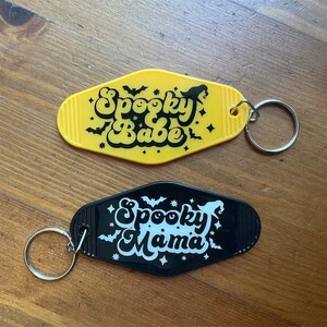 May include: Two black and yellow keychains with a retro motel key design. The yellow keychain says "Spooky Babe" in black lettering with bats and stars. The black keychain says "Spooky Mama" in white lettering with bats and stars.