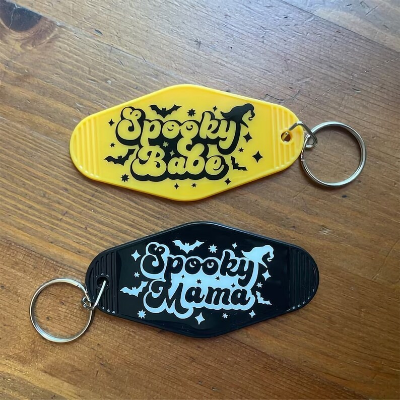 May include: Two black and yellow keychains with a retro motel key design. The yellow keychain says "Spooky Babe" in black lettering with bats and stars. The black keychain says "Spooky Mama" in white lettering with bats and stars.