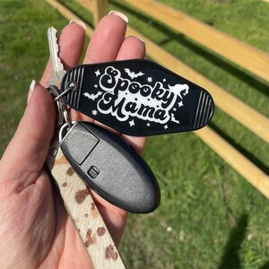 May include: A black key chain with a white "Spooky Mama" design. The keychain is attached to a brown and white spotted strap.