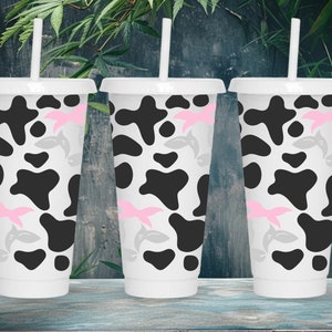 Puede incluir: Tres vasos reutilizables blancos con estampado de vaca en blanco y negro y diseños de delfines rosas y grises. Los vasos tienen pajitas de plástico transparentes.
