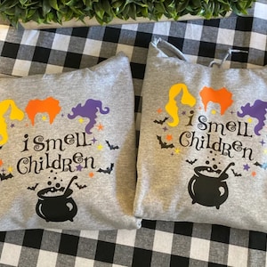 May include: Two gray hooded sweatshirts with a Halloween theme. The sweatshirts feature a cauldron, three witches, bats, and stars. The text on the sweatshirts reads "I smell children".