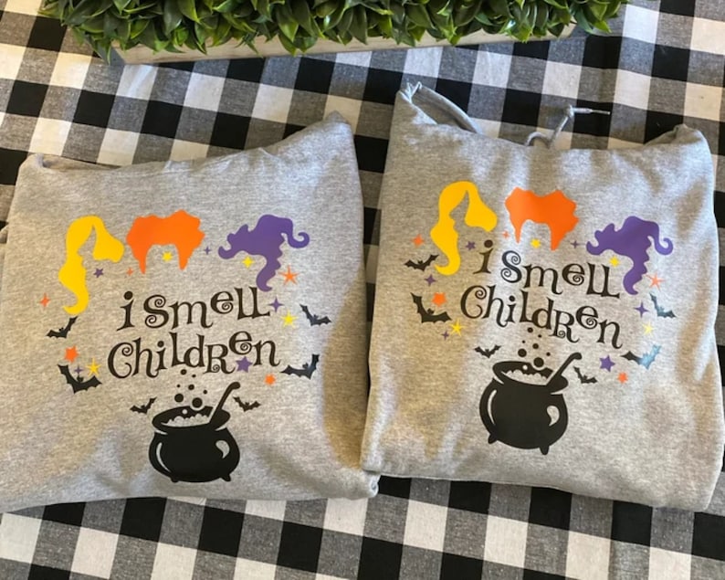 May include: Two gray hooded sweatshirts with a Halloween theme. The sweatshirts feature a cauldron, three witches, bats, and stars. The text on the sweatshirts reads "I smell children".