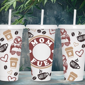 May include: Three white tumblers with lids and straws. The tumblers have different designs. One has a brown circle with the text "MOM FUEL" inside. Another has a design with coffee cups, coffee beans, hearts, and the text "WAKE UP". The third tumbler has a similar design with a woman's face and the text "WAKE UP".