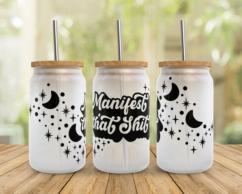K&ouml;nnte beinhalten: Drei wei&szlig;e Becher mit schwarzen Designs. Der mittlere Becher hat den Text "Manifest that Shit" in einer schwarzen, stilisierten Schrift. Die beiden anderen Becher haben ein Design mit einem Halbmond und Sternen. Jeder Becher hat einen Bambusdeckel und einen Edelstahlstrohhalm.