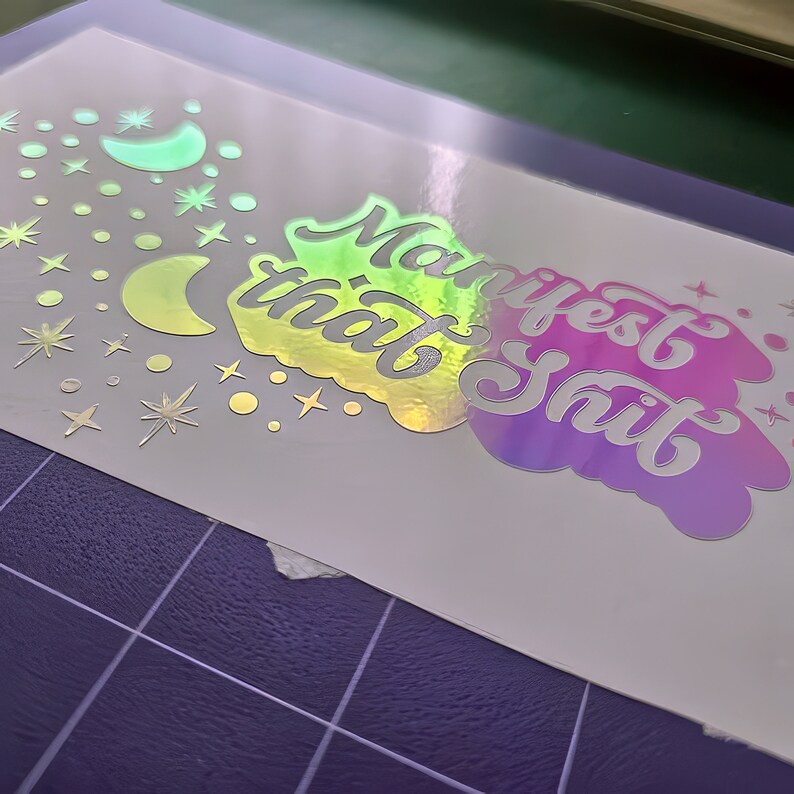 K&ouml;nnte beinhalten: Ein holografischer Aufkleber mit dem Text "Manifest that Shit" in einem Regenbogen-Farbschema. Der Aufkleber ist von silbernen Sternen und Monden umgeben.